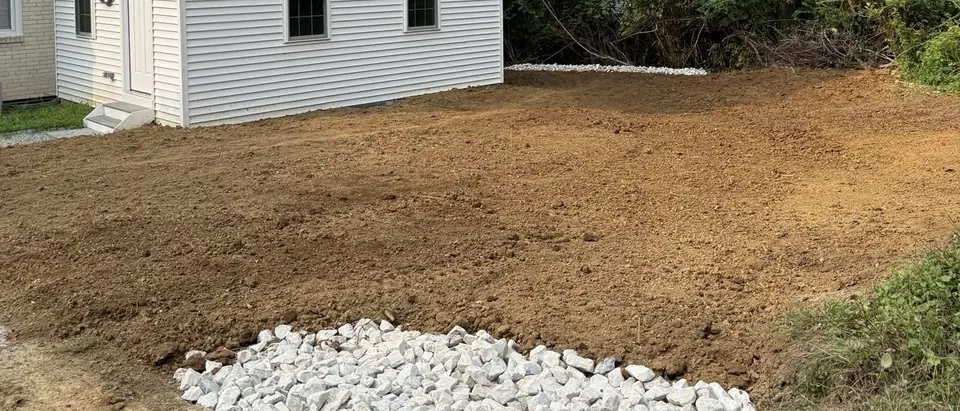 A backyard of freshly graded bare soil beside a white house, with a bed of white drainage stone in the foreground