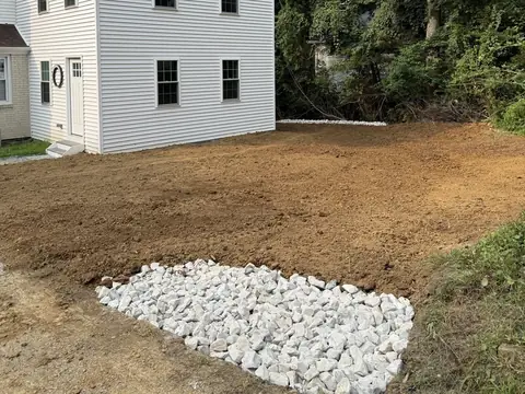 A backyard of freshly graded bare soil beside a white house, with a bed of white drainage stone in the foreground