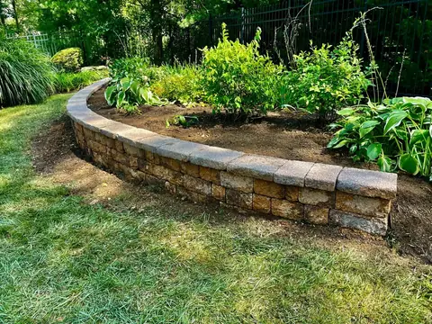 A low curved wall of tumbled block with a cap course, holding a bed of hostas and shrubs beside a lawn