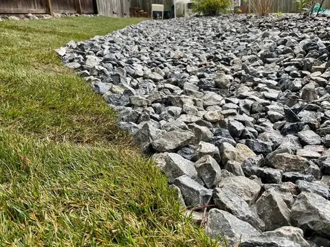 A band of angular gray stone laid along the edge of a lawn, with a wooden fence behind it