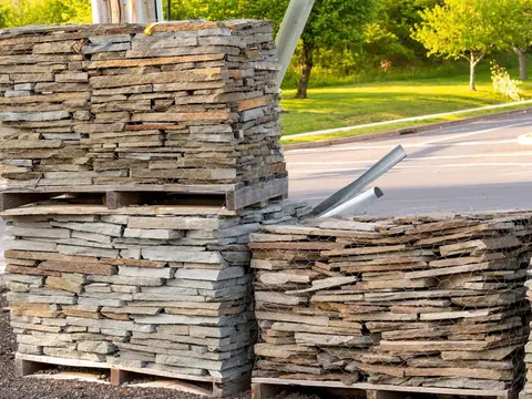 Flat pieces of natural stone stacked on wooden pallets and wrapped in wire mesh, beside a road