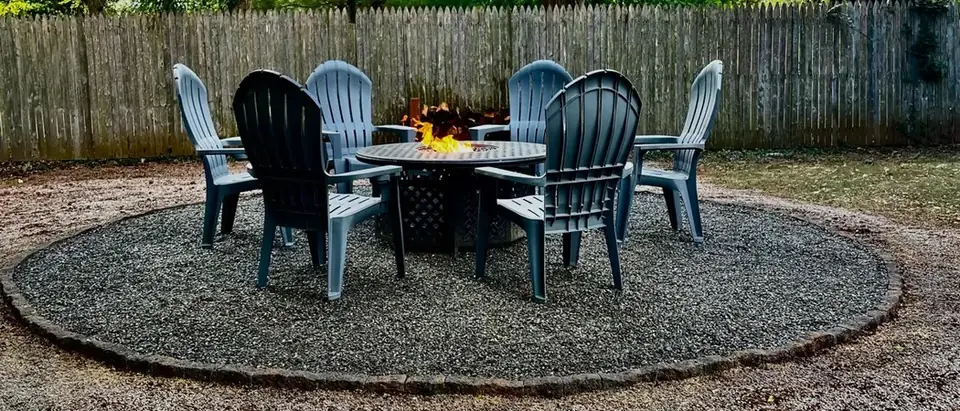 A lit fire pit table ringed by six chairs on a round gravel patio with a cobblestone edge, under trees