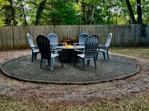 A lit fire pit table ringed by six chairs on a round gravel patio with a cobblestone edge, under trees