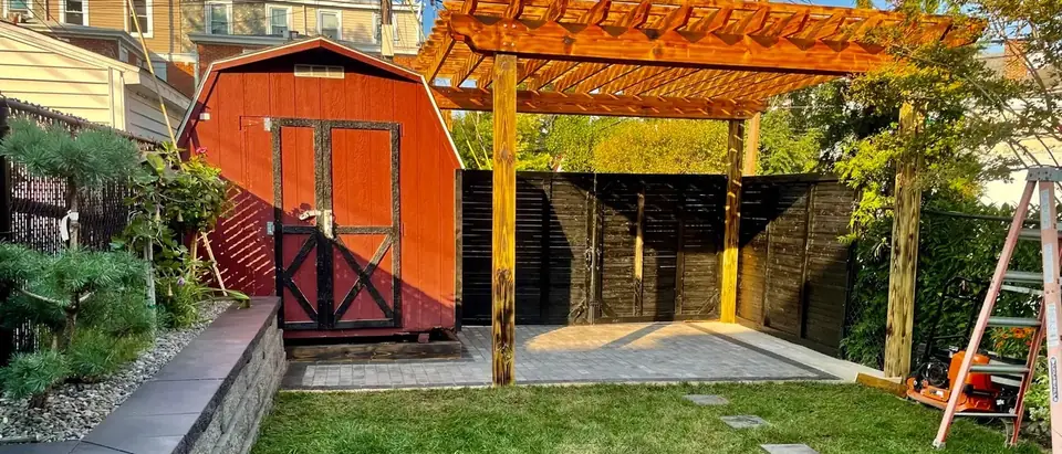 A gray paver patio under a wooden pergola beside a red shed, with a capped block planter wall and a lawn in front