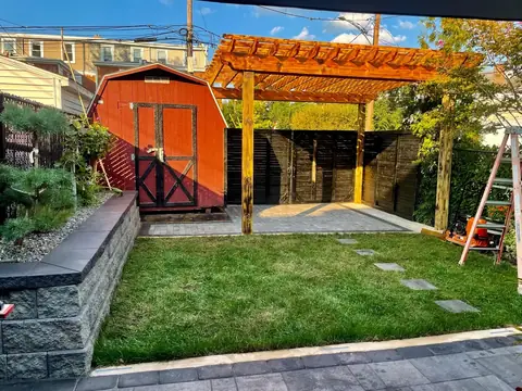 A gray paver patio under a wooden pergola beside a red shed, with a capped block planter wall and a lawn in front