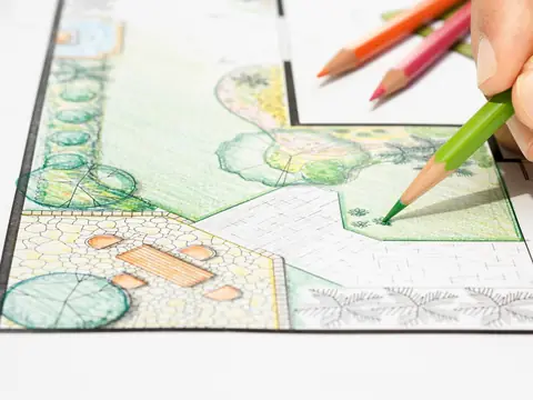 A hand coloring a garden plan on paper with a green pencil, with more colored pencils lying beside it