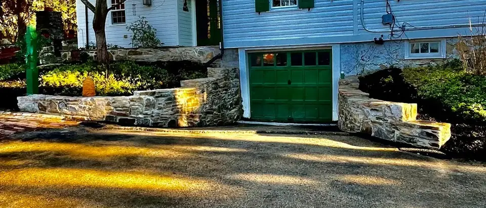 Natural fieldstone walls on both sides of a garage entrance, below a white house with green shutters