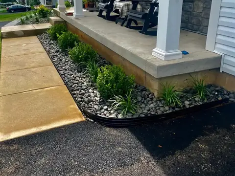A bed of river rock with boxwoods and grassy plants along the edge of a concrete porch, held by metal edging