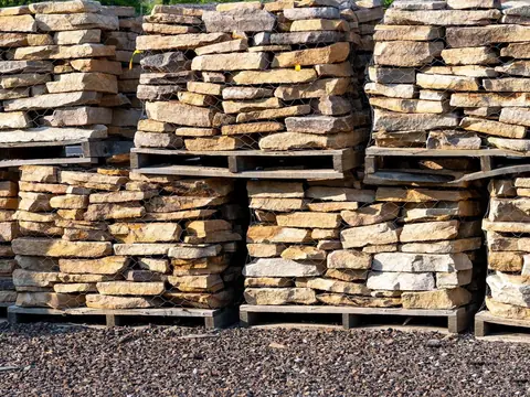 Rough tan building stone stacked on wooden pallets under wire mesh, on a gravel yard
