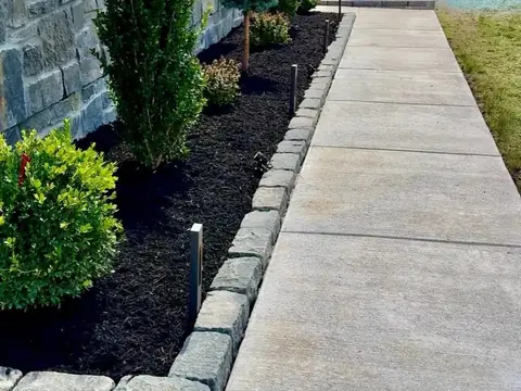 A concrete walkway edged with gray cobblestones, beside a planted bed along a stone-faced house