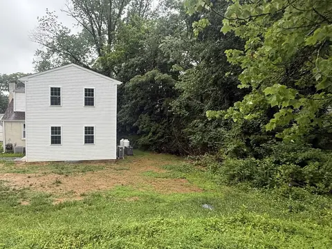 A side yard of thin grass and bare soil beside a white two-story house, at the edge of woods